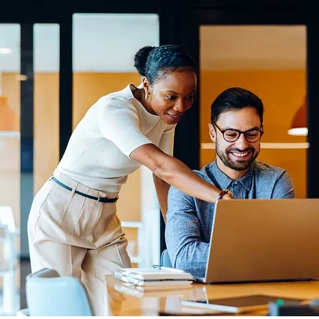 A woman standing and pointing at a laptop screen while a seated man wearing glasses smiles and looks at the laptop in a bright office.