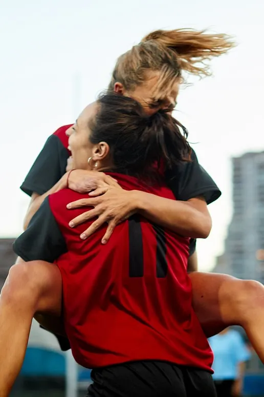 Two female soccer players in red and black jerseys embracing joyfully on the field, with one player lifted off the ground.