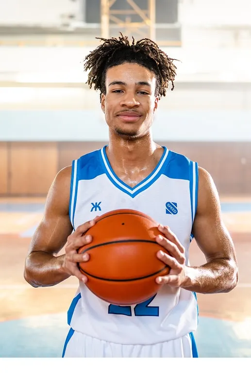 Smiling male basketball player in white and blue jersey holding a basketball in an indoor court.