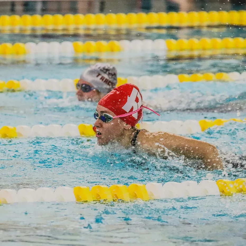 Two swimmers competing in a swimming pool, with one in a red swim cap and goggles in the foreground.