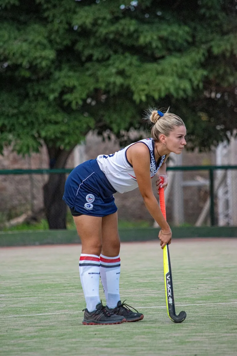 Female field hockey player in white and blue uniform bent forward holding a hockey stick on a green field.