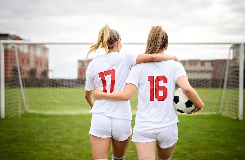 Two female soccer players in white uniforms with numbers 17 and 16 walking arm in arm toward a goalpost, one holding a soccer ball.
