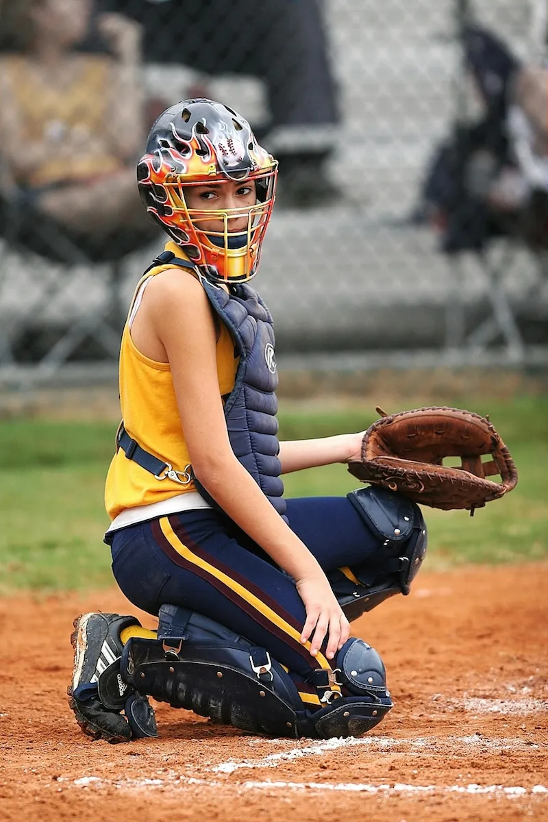 Youth softball catcher wearing protective gear and a flame-designed helmet crouches behind home plate ready to catch a ball.