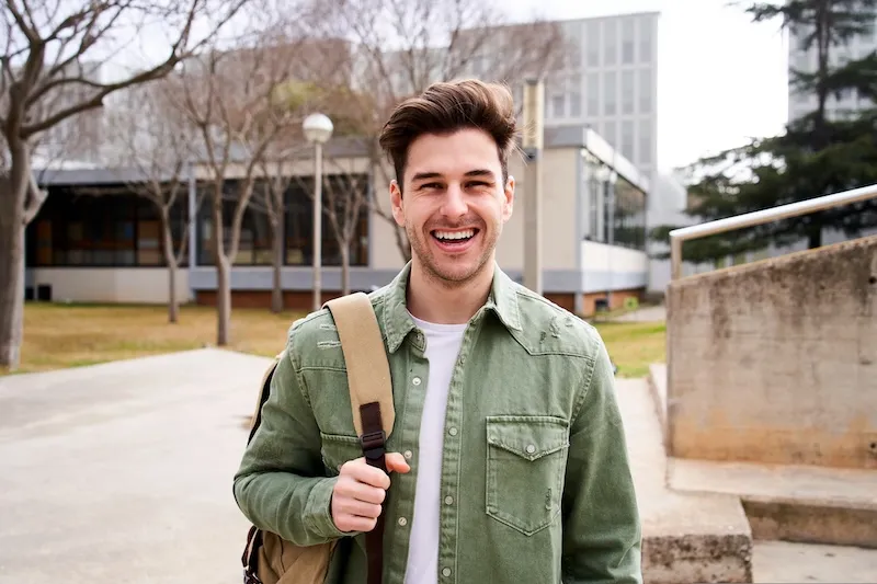 Smiling young man wearing a green jacket and carrying a backpack outdoors on a campus pathway.