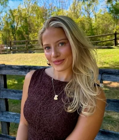 Smiling young woman with long blonde hair wearing a dark sleeveless top and pendant necklace sitting on a wooden bench outdoors with trees and grass in the background.