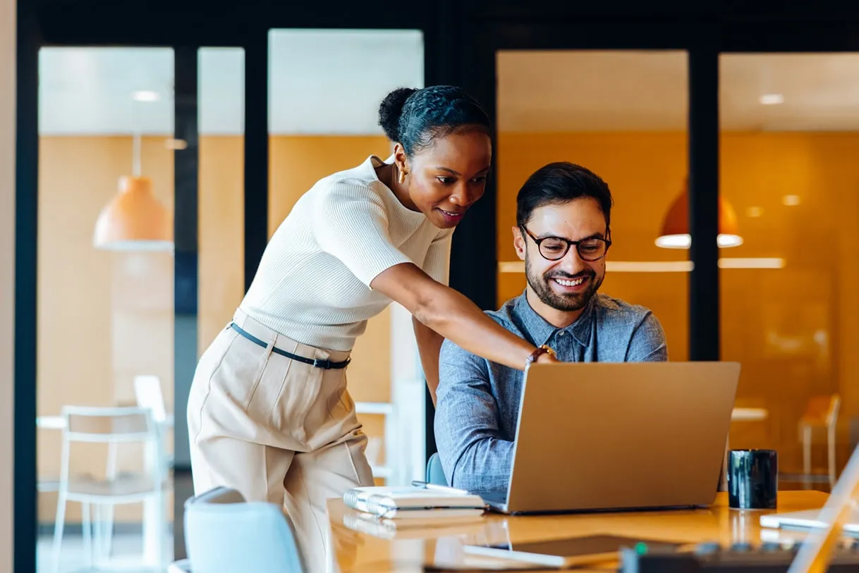 Smiling woman points at laptop screen while man with glasses looks on in modern office.