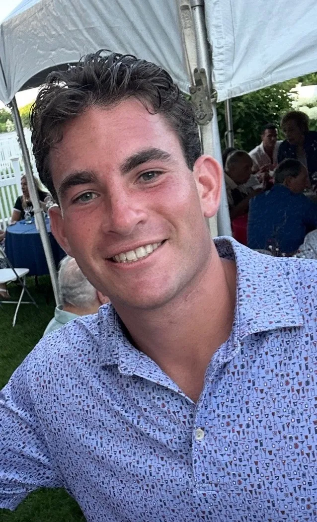 Smiling young man with curly dark hair wearing a patterned blue button-up shirt at an outdoor event under a white tent.