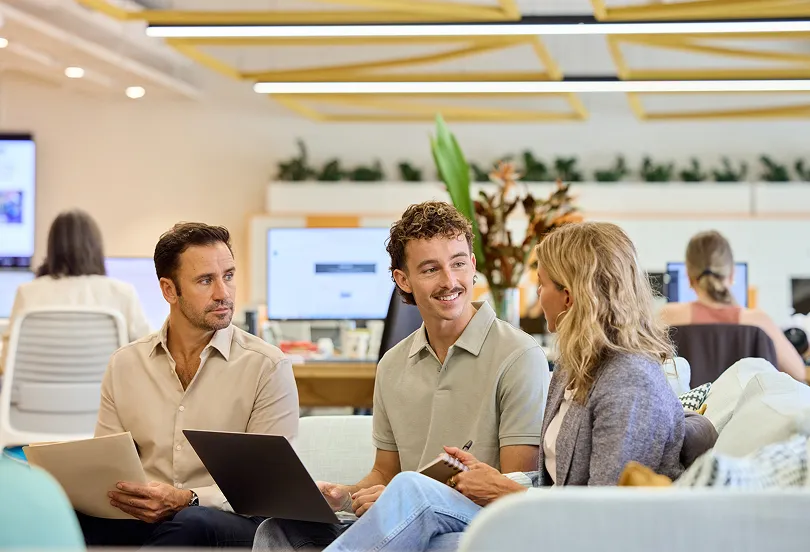 Three colleagues seated together in a bright, open office space, reviewing documents and a laptop while discussing ideas collaboratively.