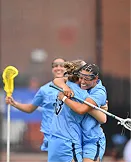 Two female lacrosse players in blue uniforms smiling and celebrating on the field.