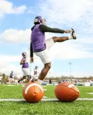 American football player in purple kicking ball on field while another watches, under blue sky with clouds.