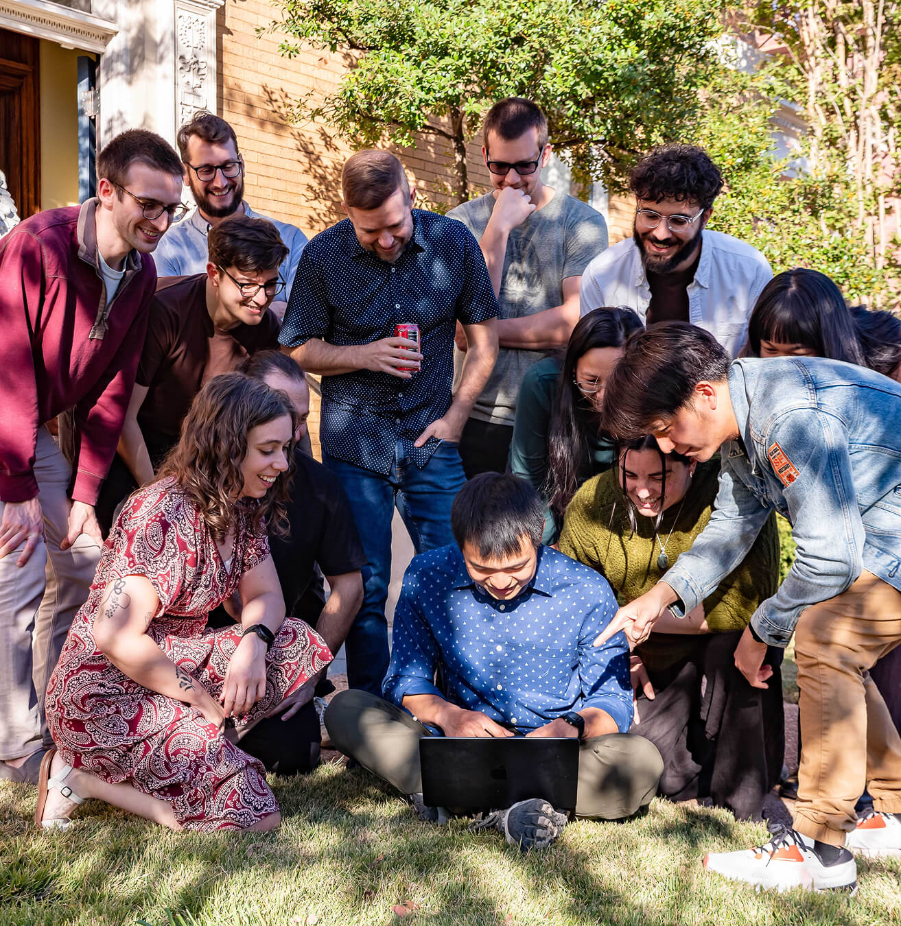Group of diverse young adults gathered outside on grass looking at a laptop, smiling and engaged.