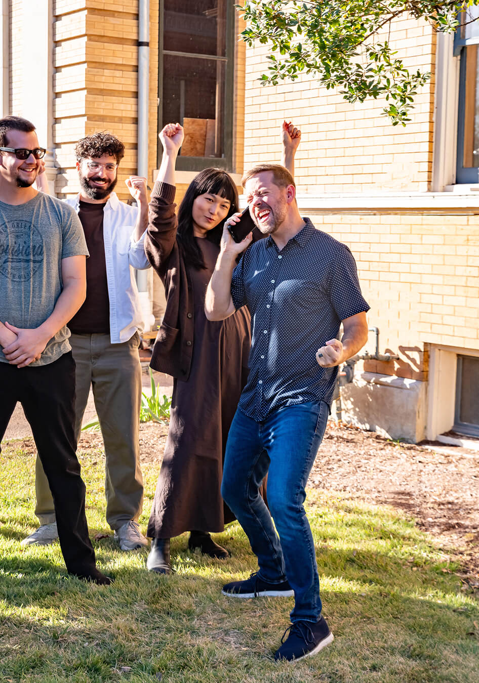 Group of four diverse people standing outside a brick building, celebrating with raised fists and a man excitedly talking on a phone.