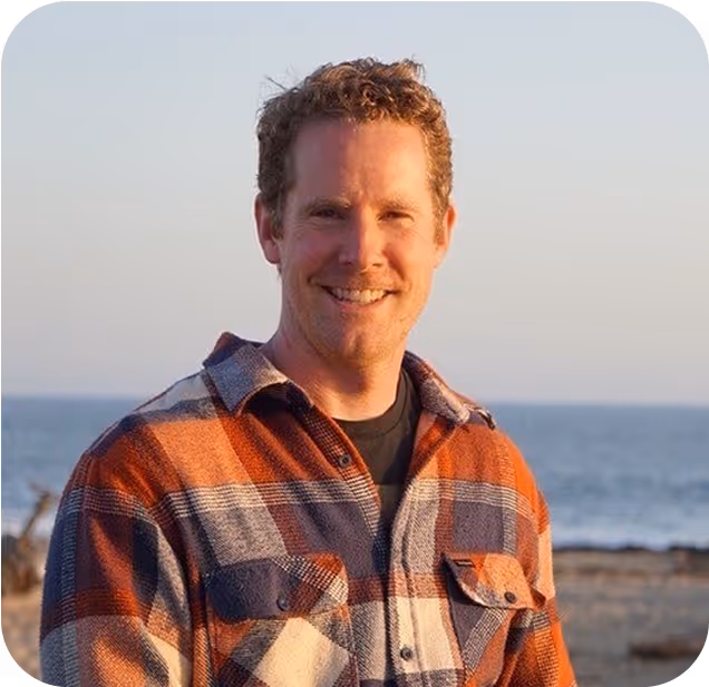 Smiling person in plaid shirt standing by ocean at sunset
