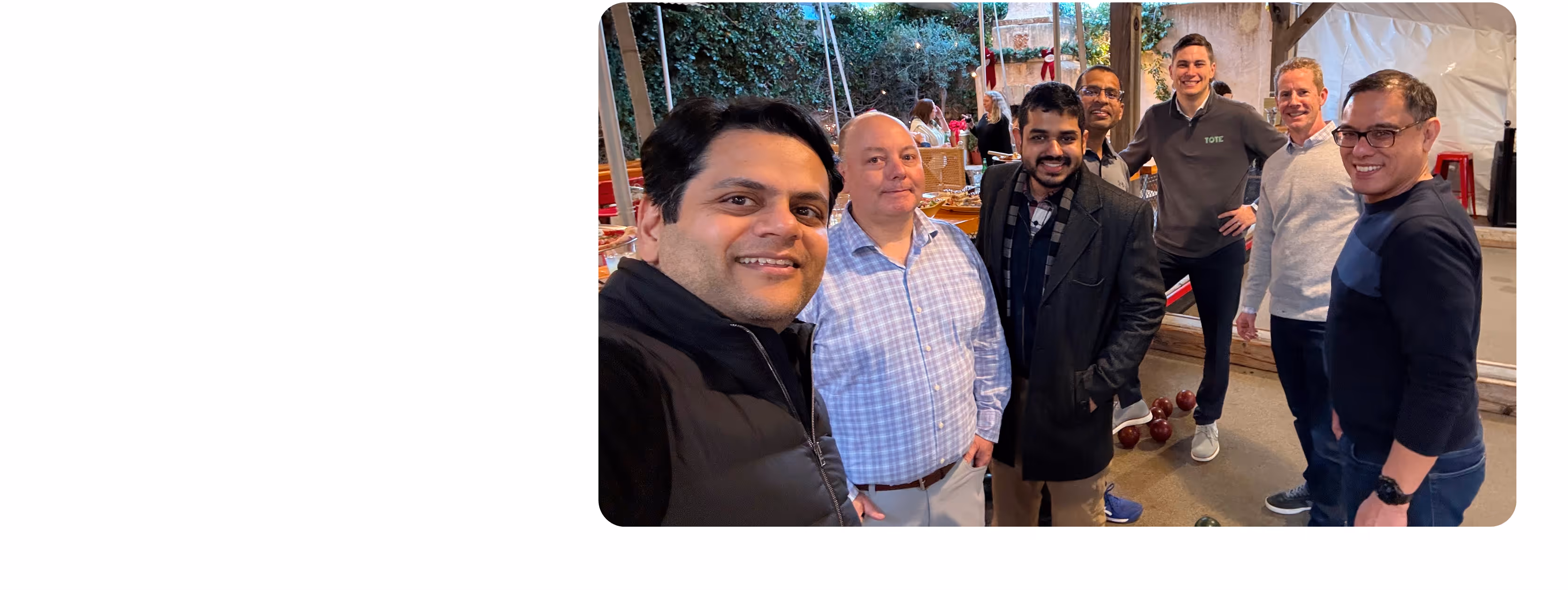 Seven men smiling and posing for a group selfie indoors with bocce balls on the floor nearby.
