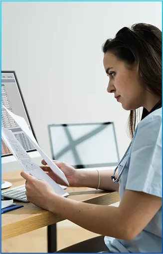 A healthcare professional reviews documents while sitting at a desk, with a computer monitor displaying data in the background.
