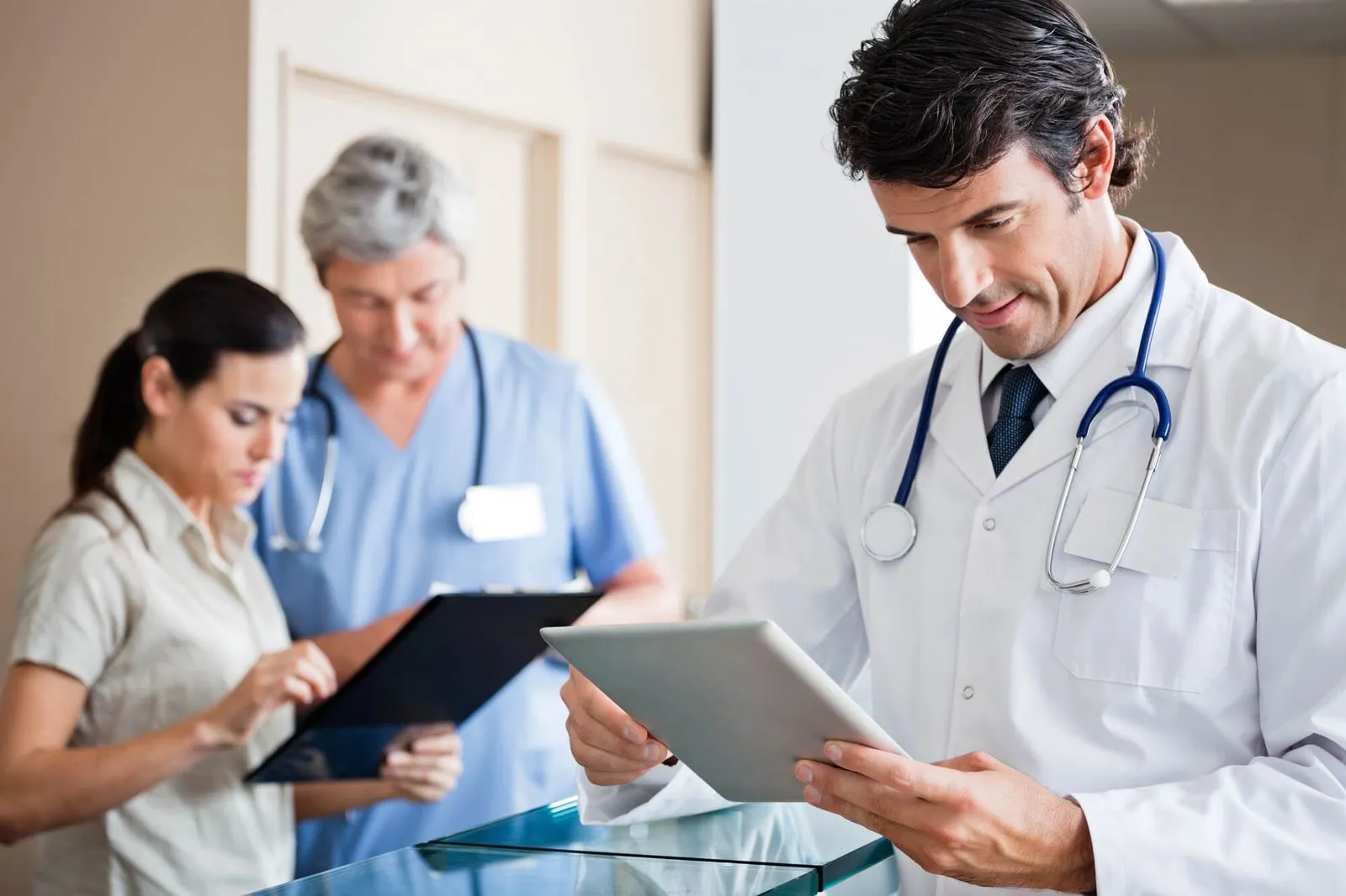 A doctor in a white coat using a tablet, with two colleagues in scrubs discussing medical documents in a modern clinic.
