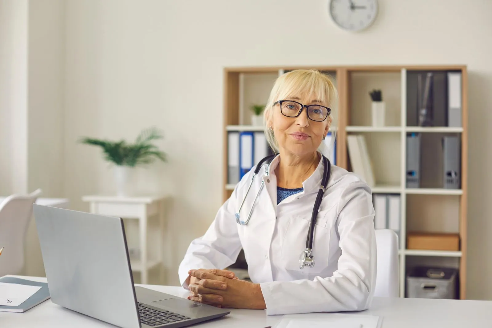 A healthcare professional in a white coat sits at a desk with a laptop, surrounded by bookshelves and greenery in a modern office setting.
