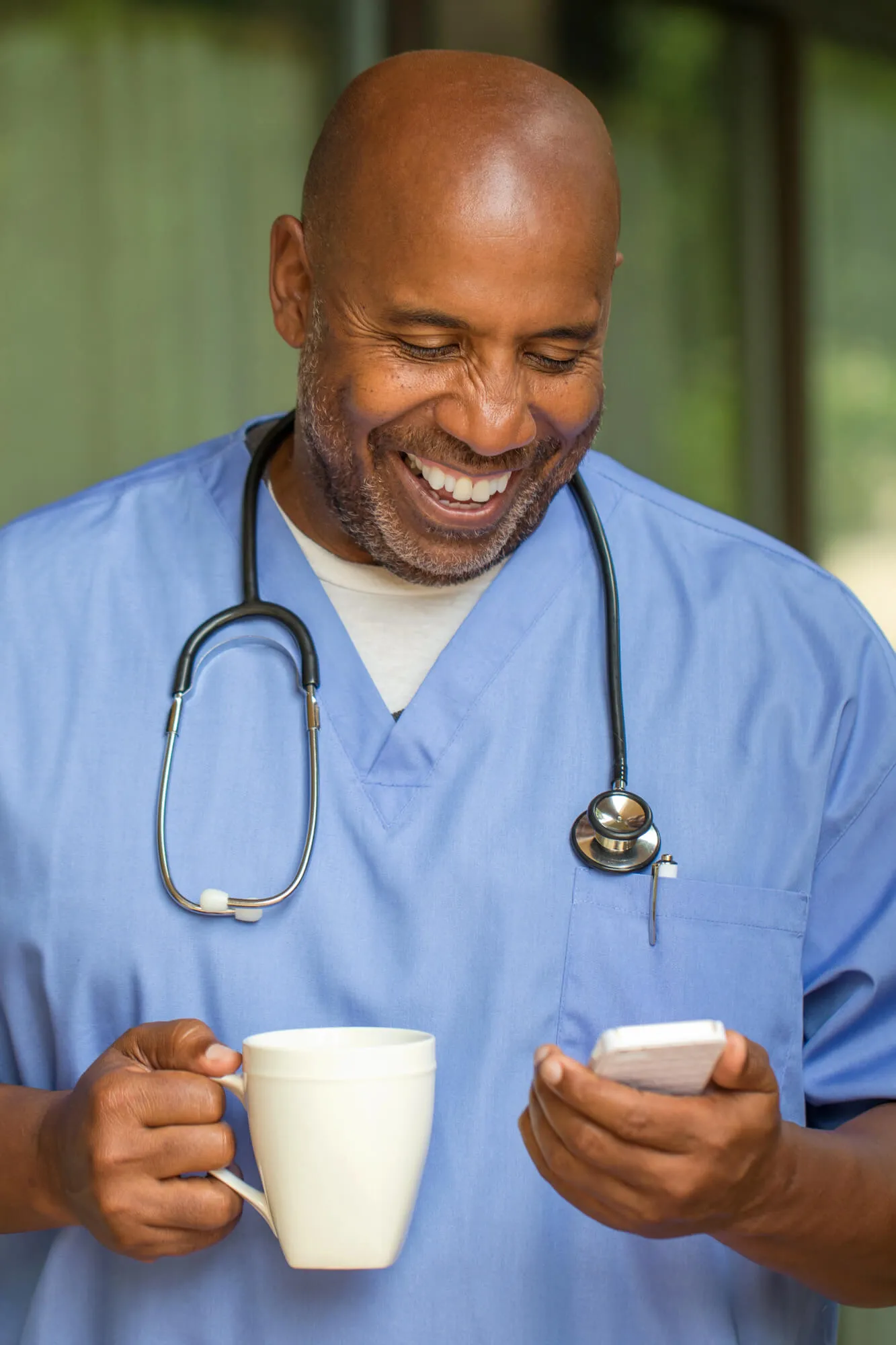 A healthcare worker in scrubs holds a white coffee mug in one hand and a smartphone in the other, standing outdoors.