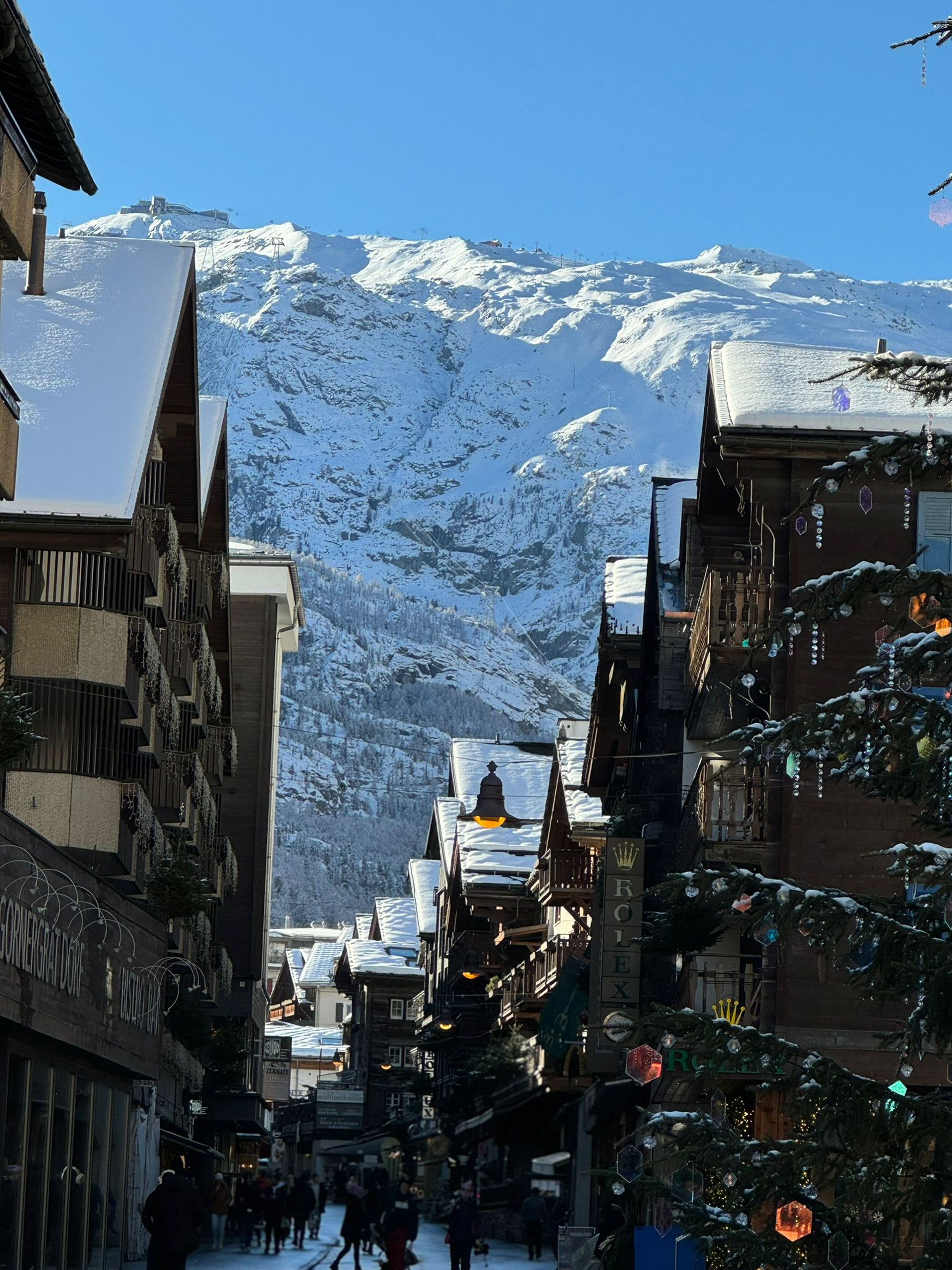 Calle de un pueblo alpino cubierta de nieve, con chalets de madera, personas caminando y una gran montaña nevada bajo un cielo azul despejado.
