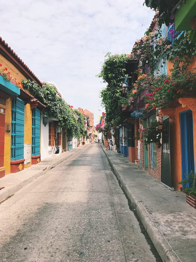 Narrow street lined with colorful buildings and vibrant flowering plants hanging over balconies under a cloudy sky.