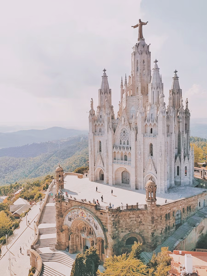 The Temple Expiatori del Sagrat Cor, a large white church with multiple spires and a statue of Jesus on top, situated on a hill with surrounding greenery and distant mountains.