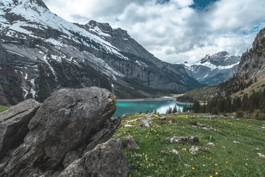 Scenic mountain landscape with snow-capped peaks, turquoise lake, green meadow with yellow wildflowers, and large foreground rocks.