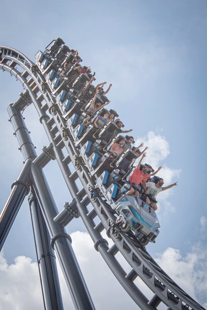 People wearing masks ride a roller coaster descending on a track against a blue sky with clouds.