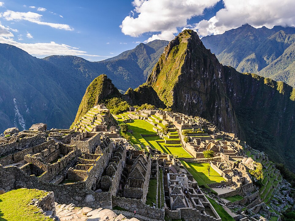 Wide view of Machu Picchu ruins with green terraces and towering mountains under a blue sky with clouds.