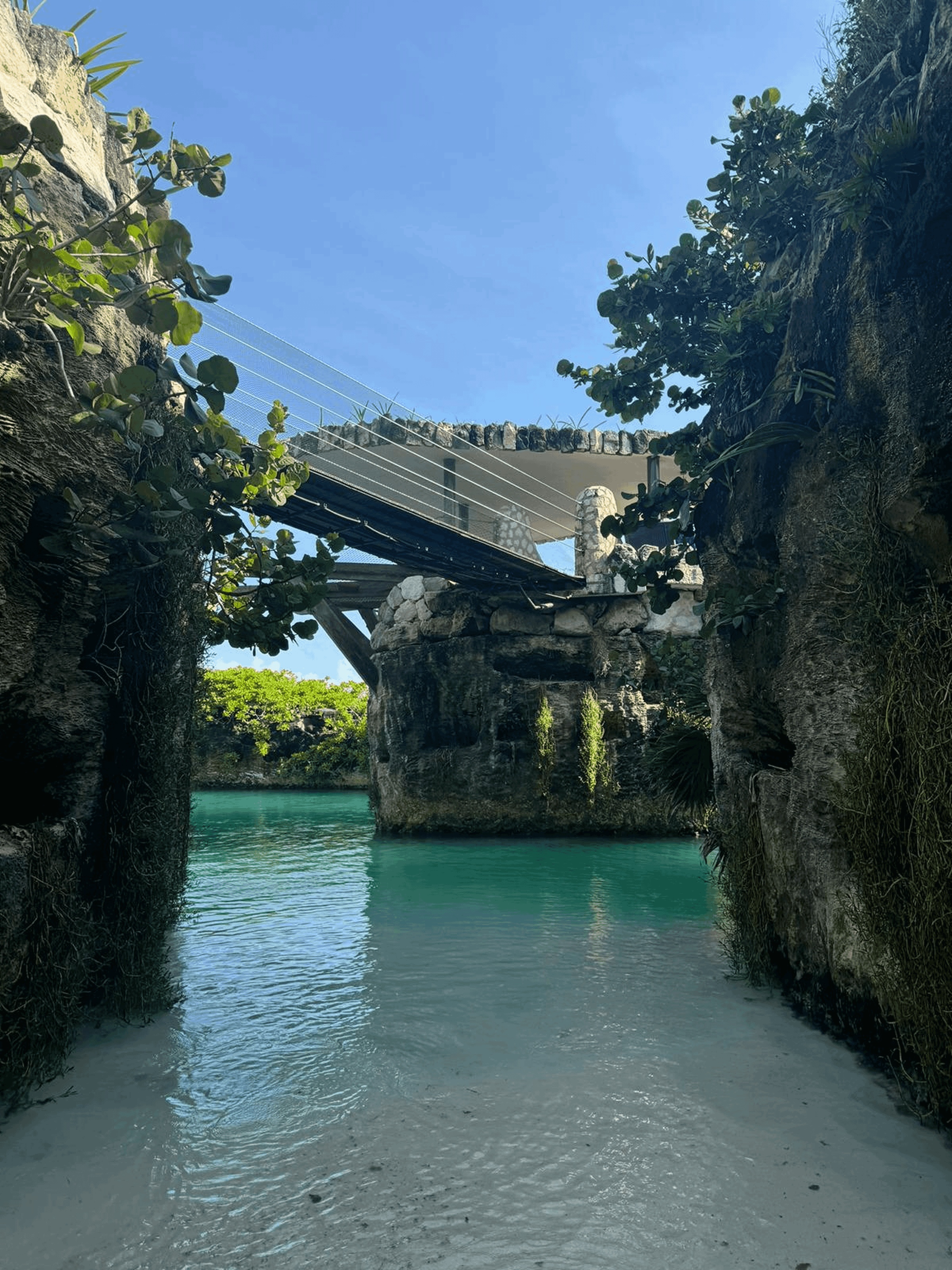 Stone bridge with cable railings over a turquoise waterway surrounded by rocky cliffs and green foliage under a clear blue sky.