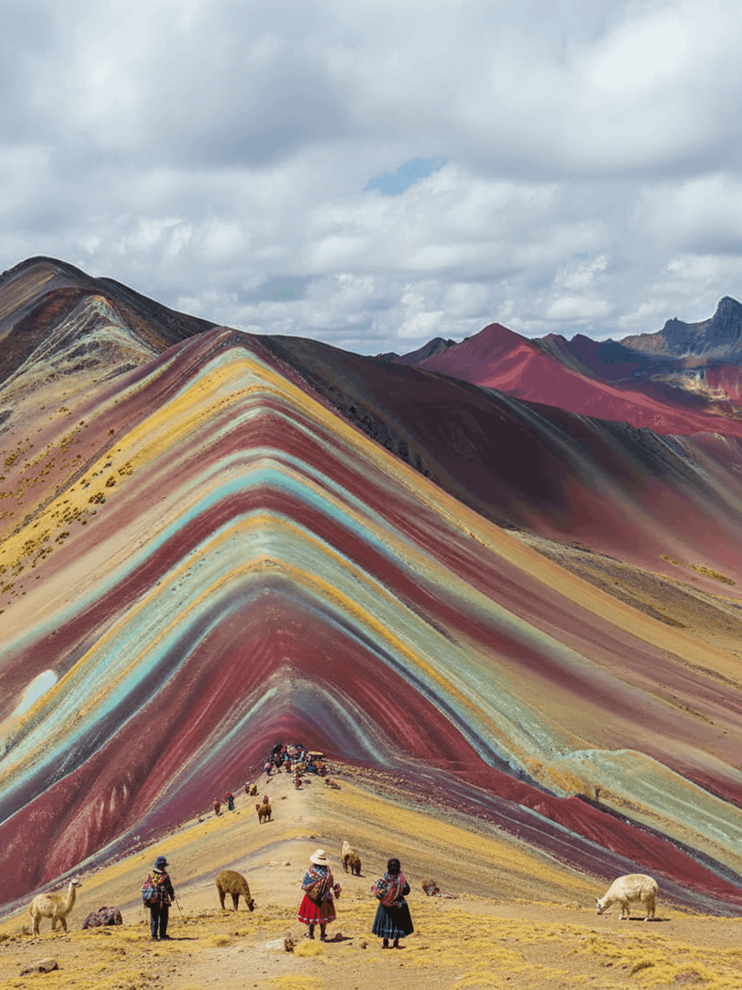 Tourists and llamas on a path near the colorful striped slopes of Rainbow Mountain under a cloudy sky.