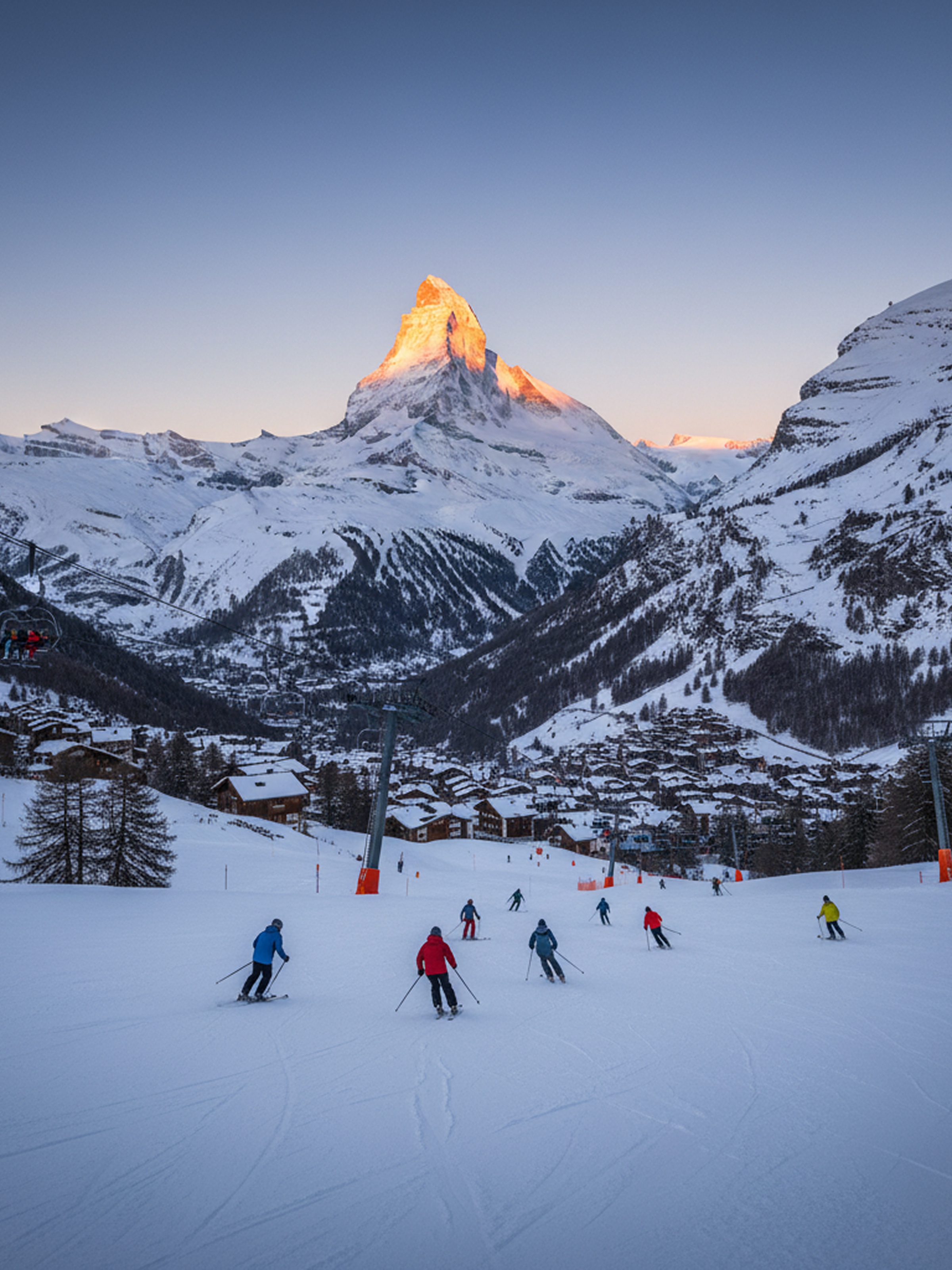 Skiers descending a snowy slope with a village below and a mountain peak bathed in sunset light.