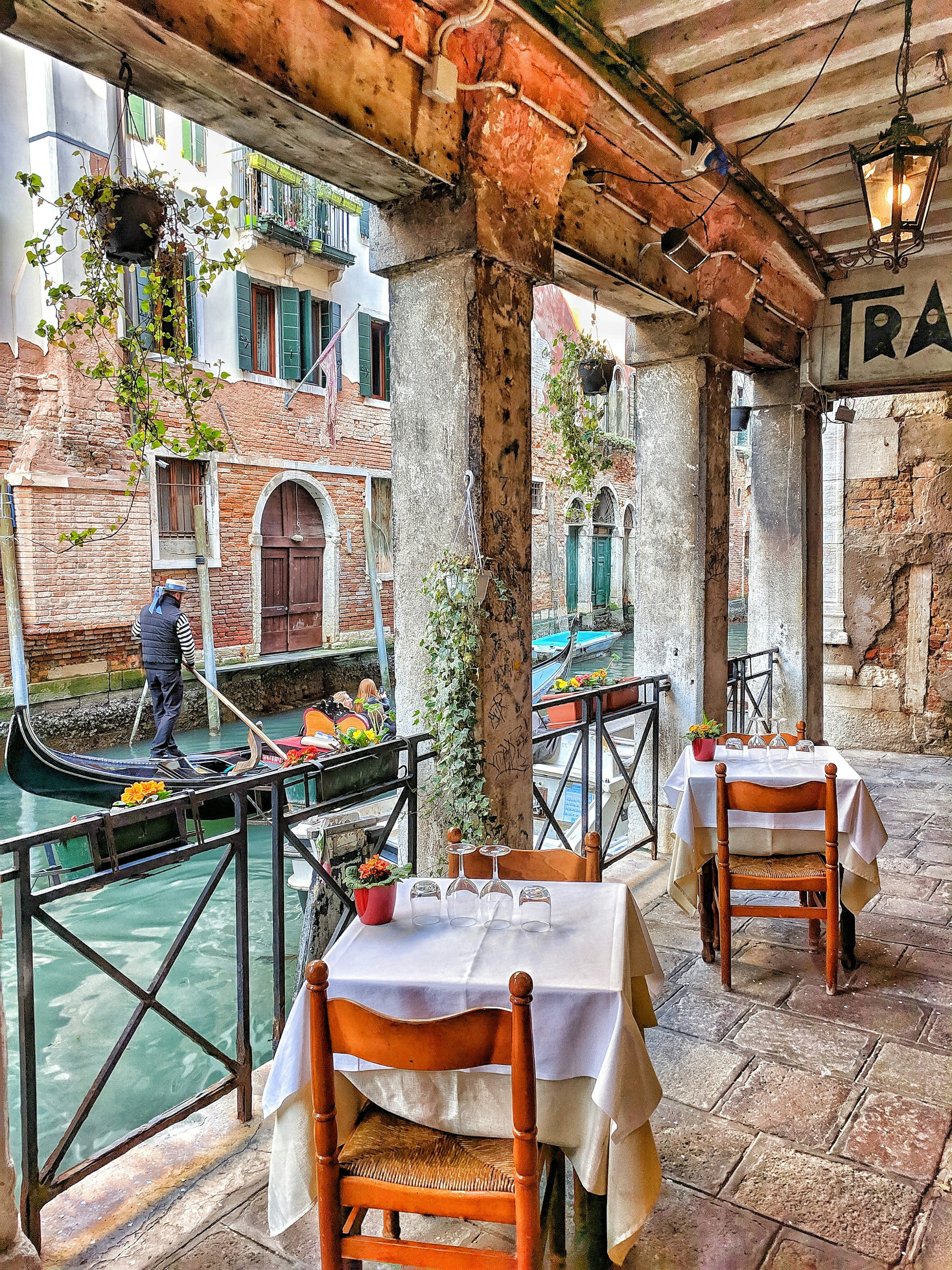 Outdoor restaurant seating by a Venetian canal with gondola and historic brick buildings in the background.