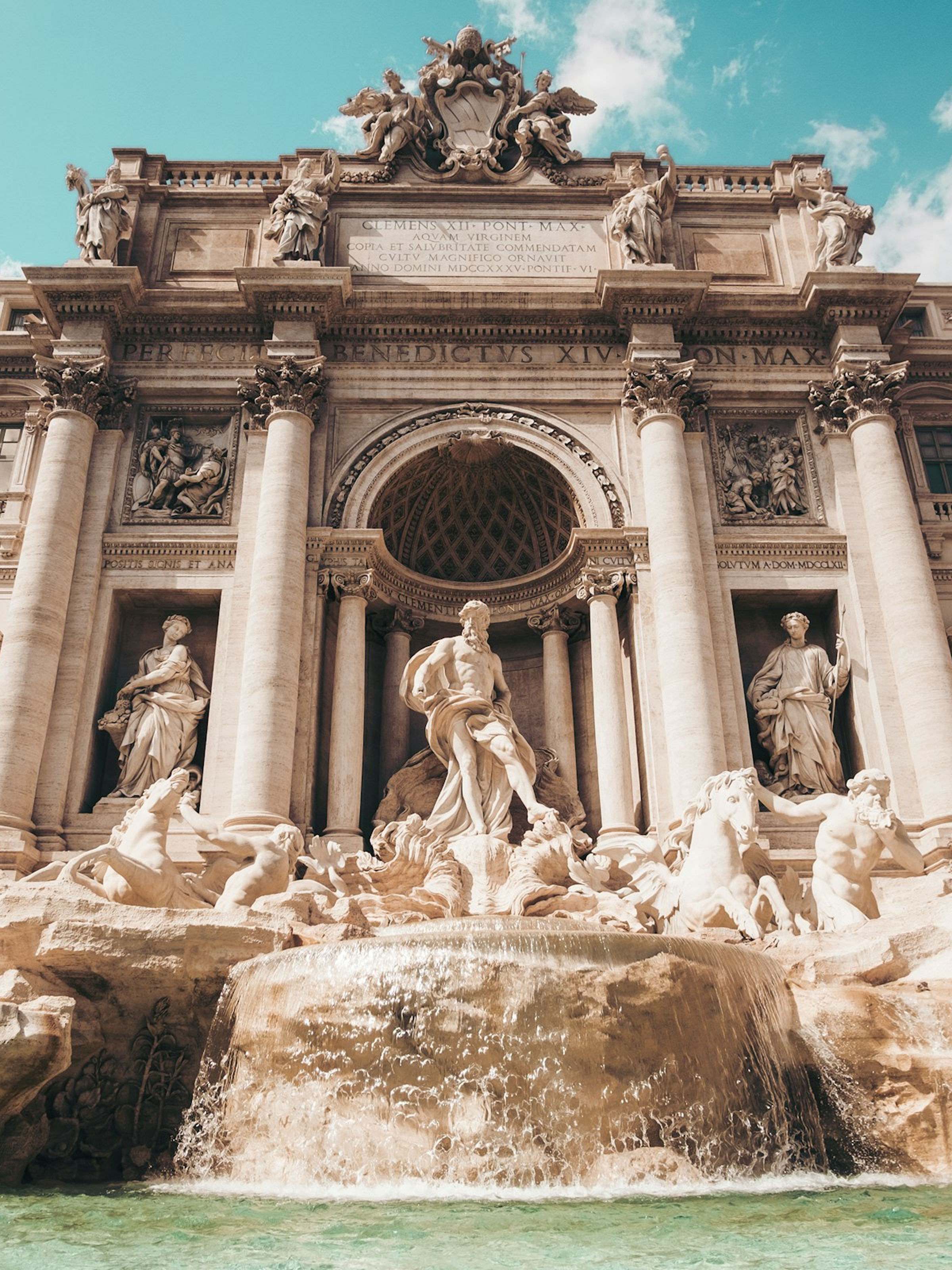 Front view of the Baroque Trevi Fountain with statues and cascading water under a blue sky.