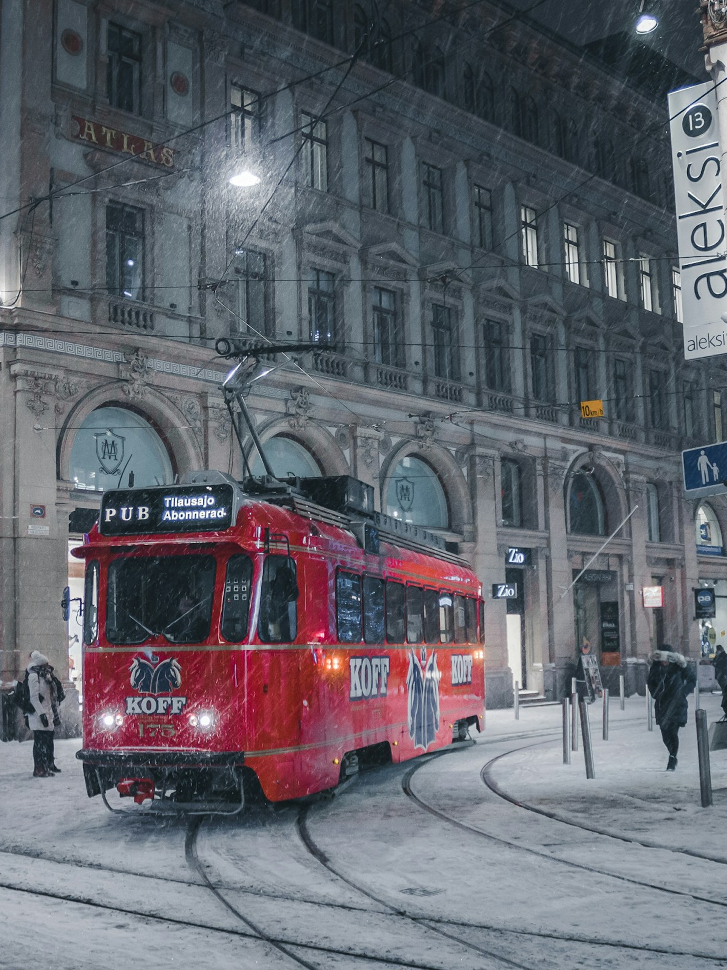 Red tram labeled KOFF traveling on snowy street in front of historic building during nighttime.