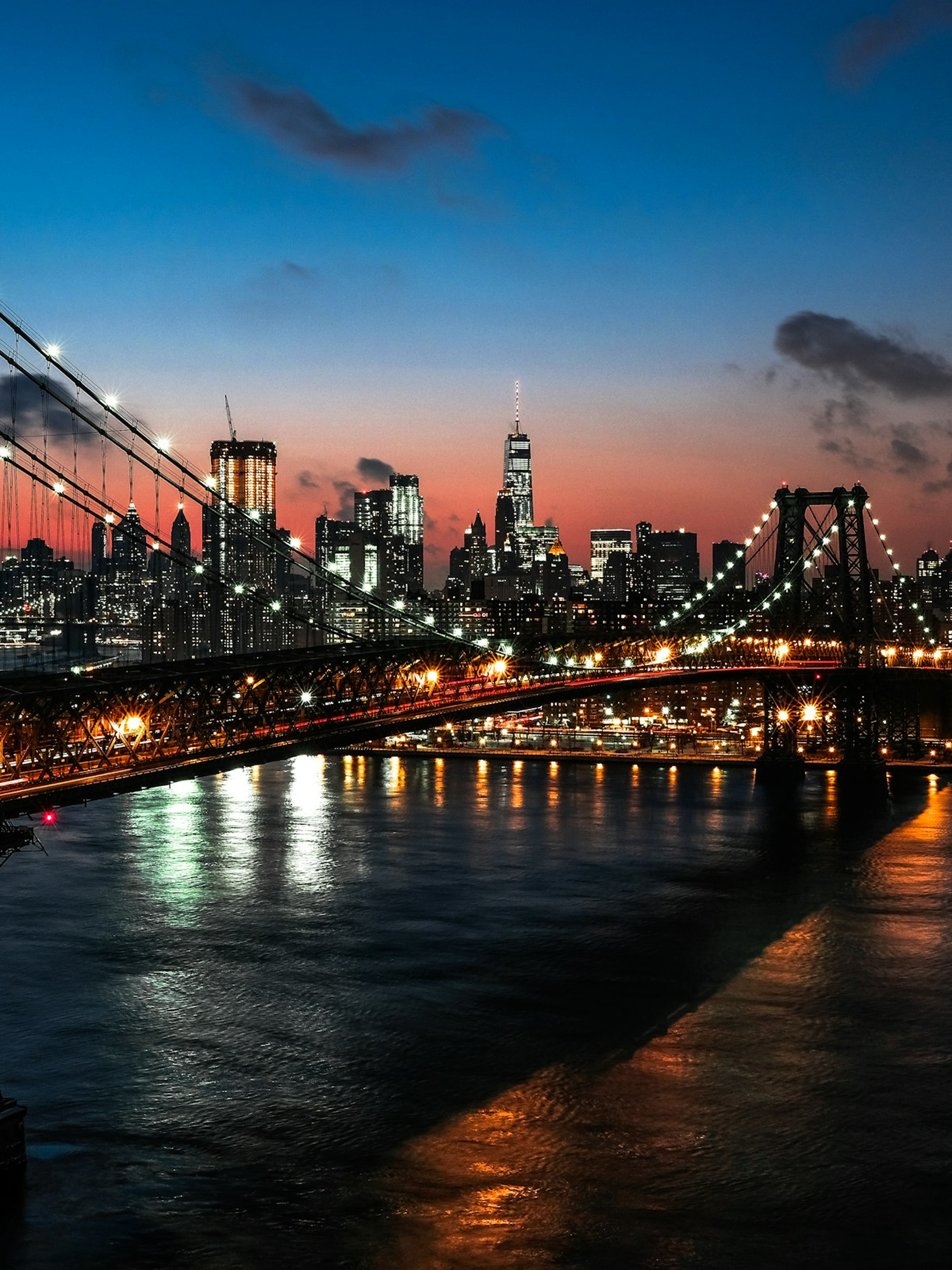 Illuminated suspension bridge over a river with a city skyline at sunset and a clear blue to orange gradient sky.