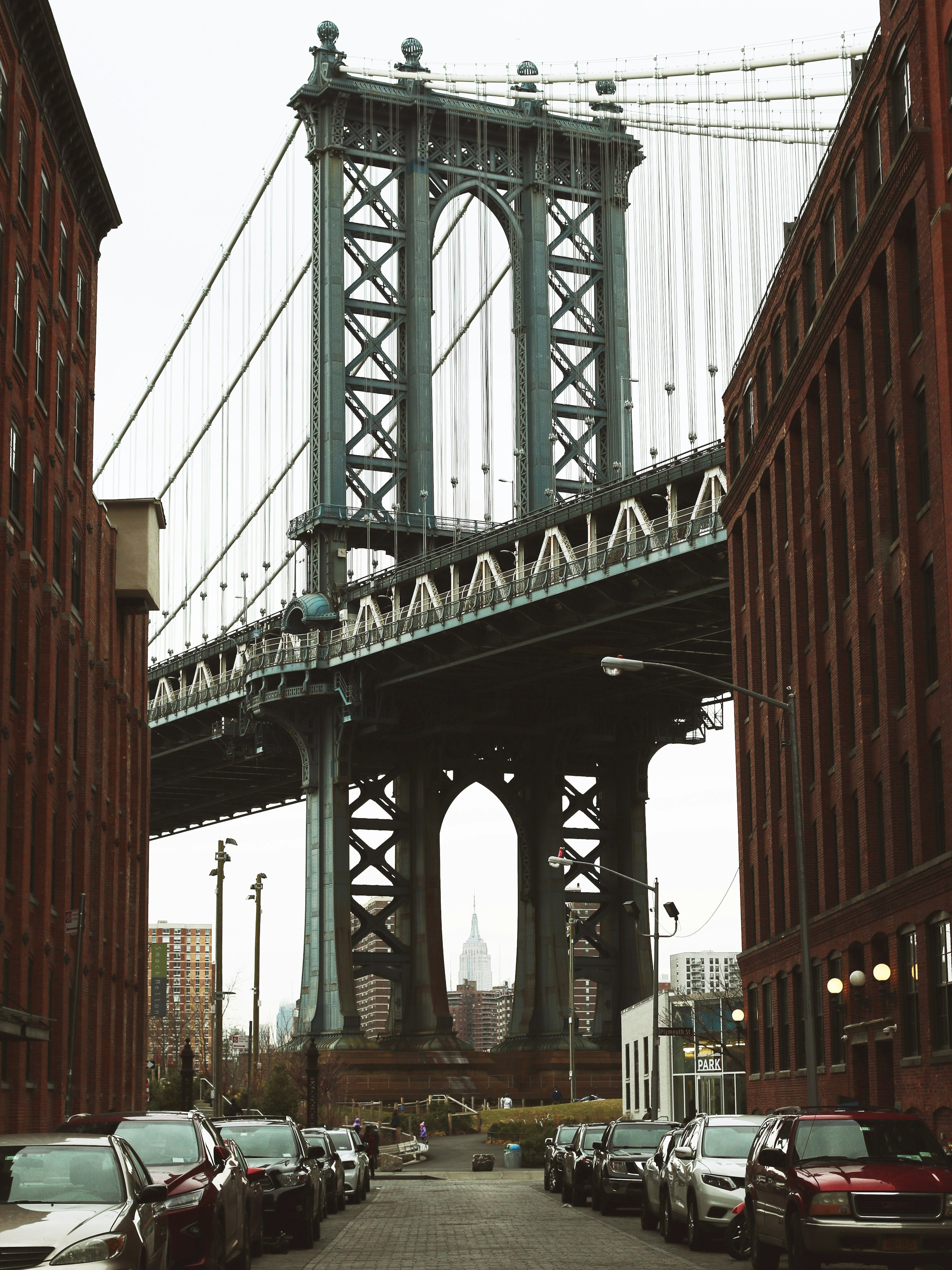 View of the Manhattan Bridge framed by brick buildings with parked cars along the street and the Empire State Building visible through the bridge arches in the background.