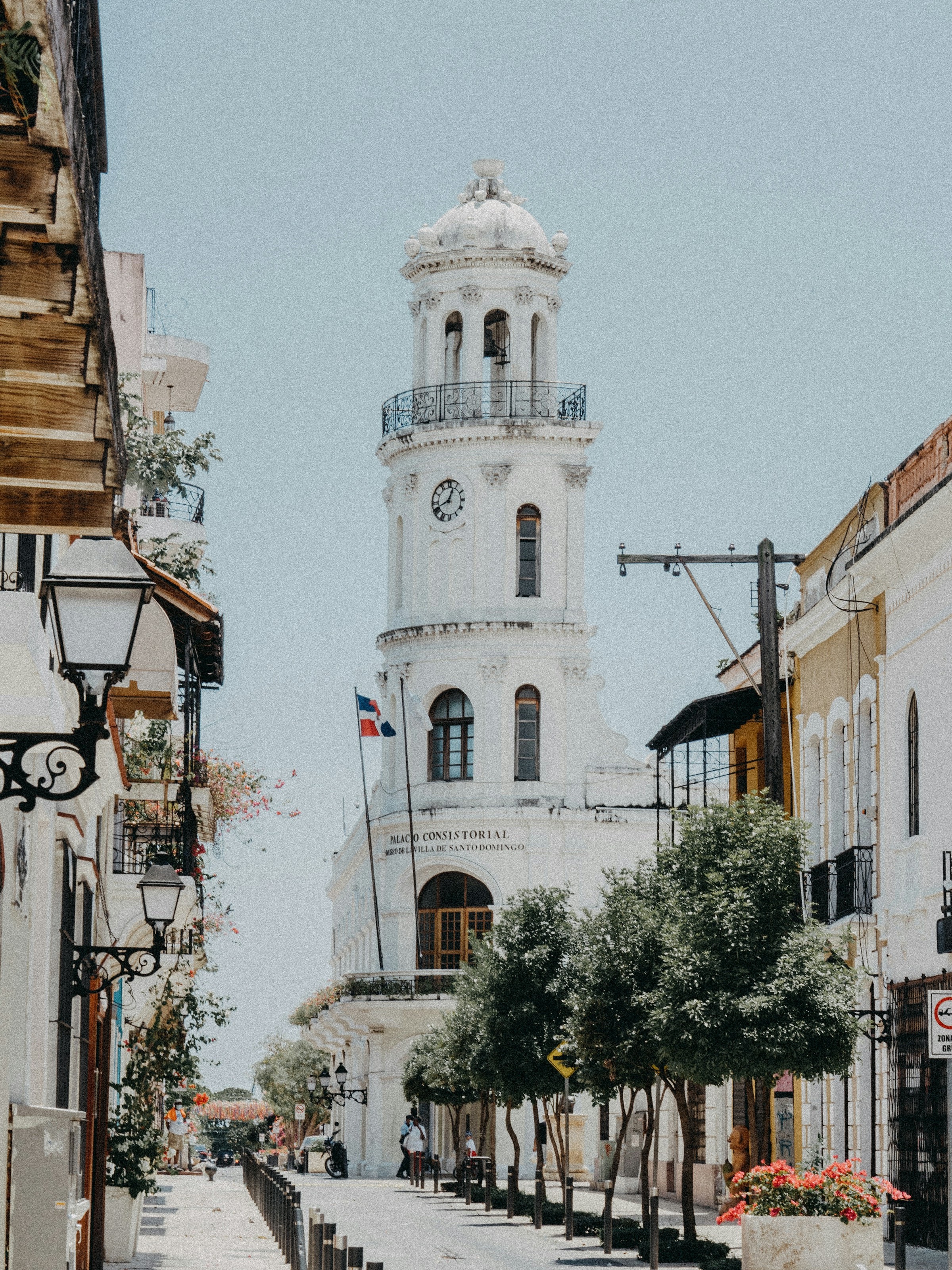 White historic building with clock tower and Dominican Republic flag along a street lined with trees and lampposts.