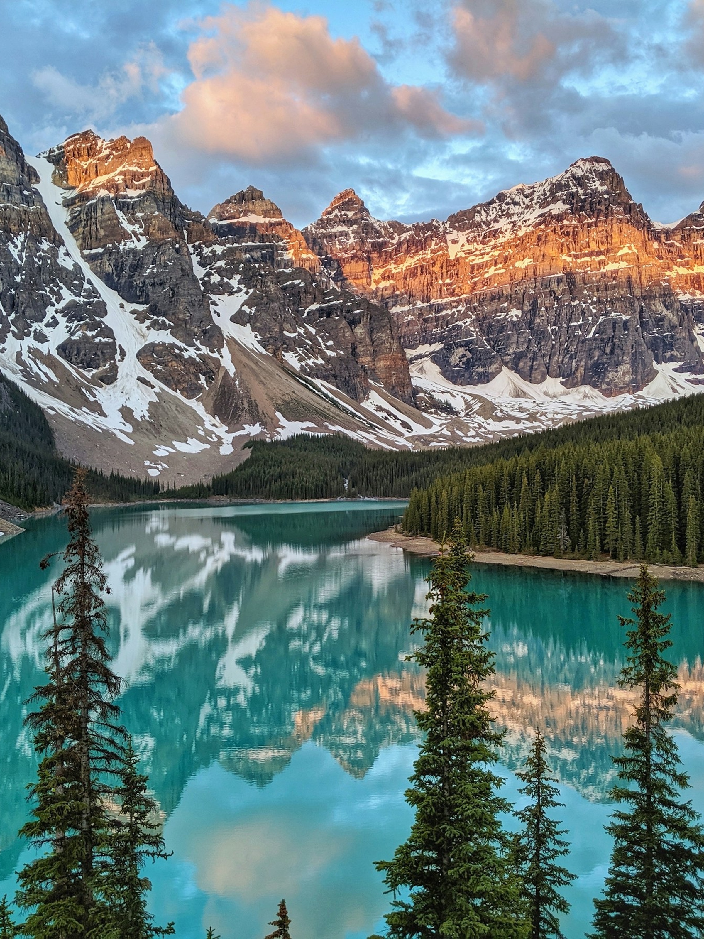 Turquoise lake reflecting snow-capped mountains and pine trees at sunset.