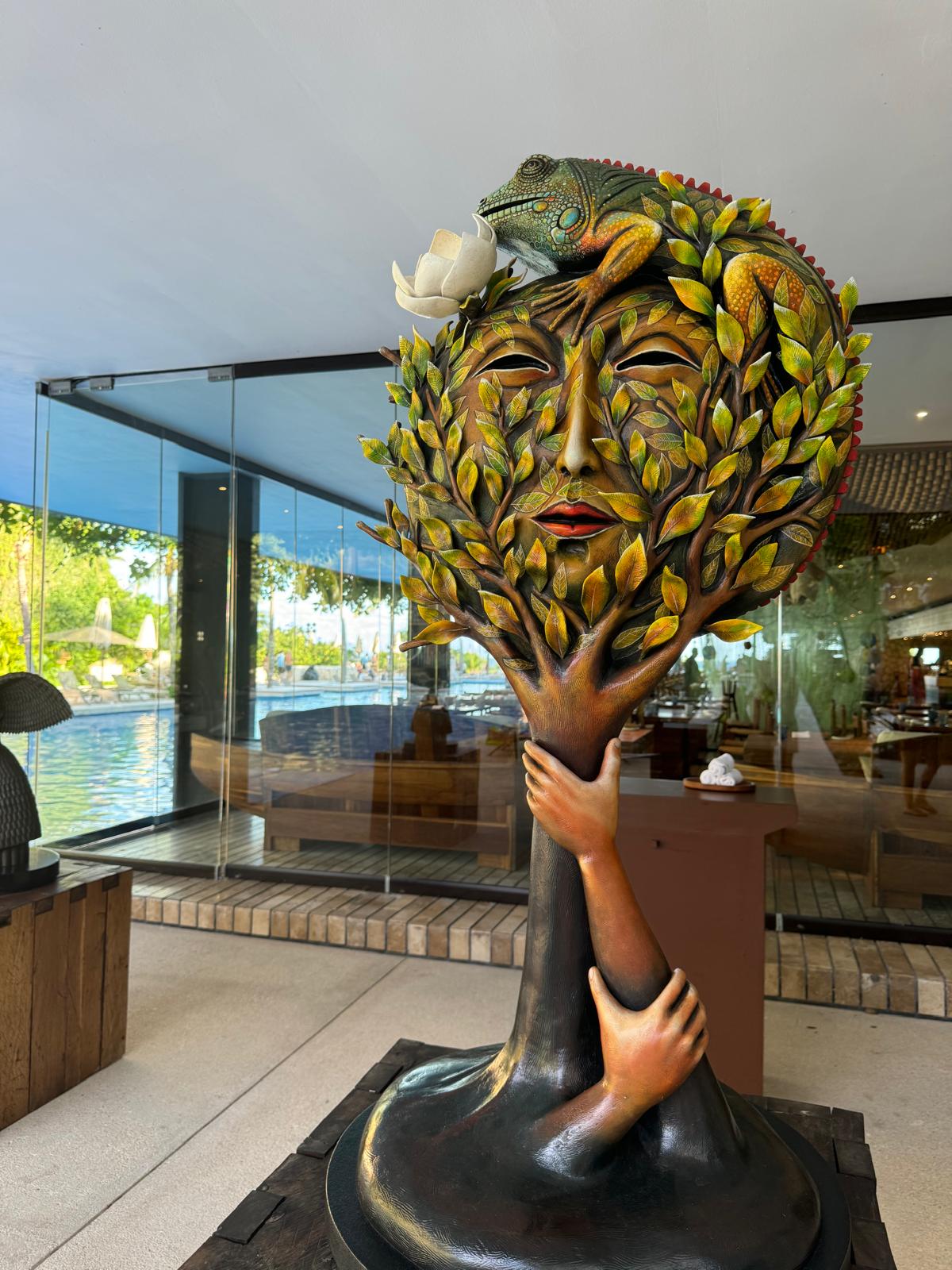 Artistic tree sculpture with human facial features, two hands hugging the trunk, and a colorful lizard perched on top holding a white flower.