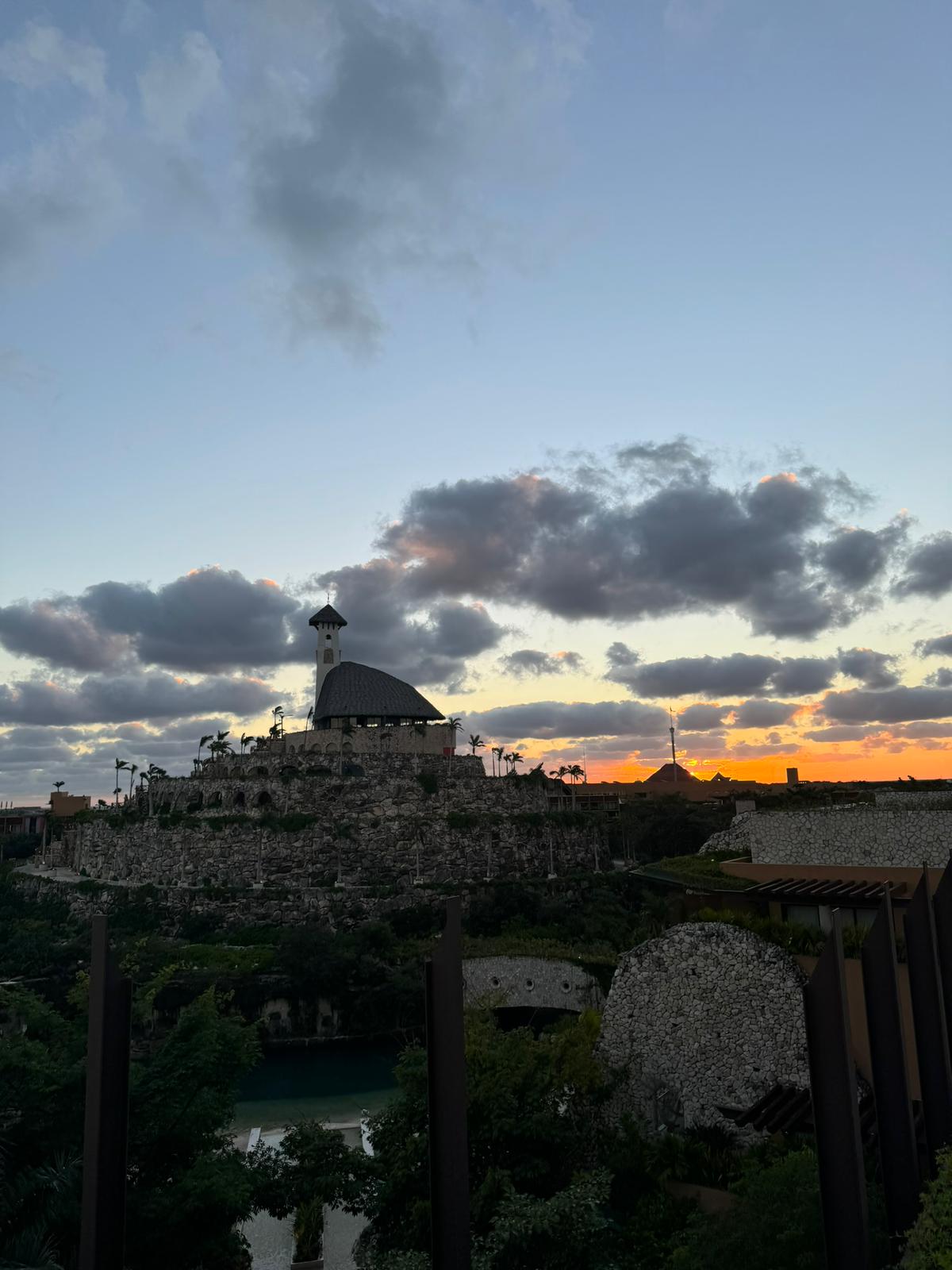 Stone building with a tower on a hill surrounded by vegetation during a sunset with scattered clouds.
