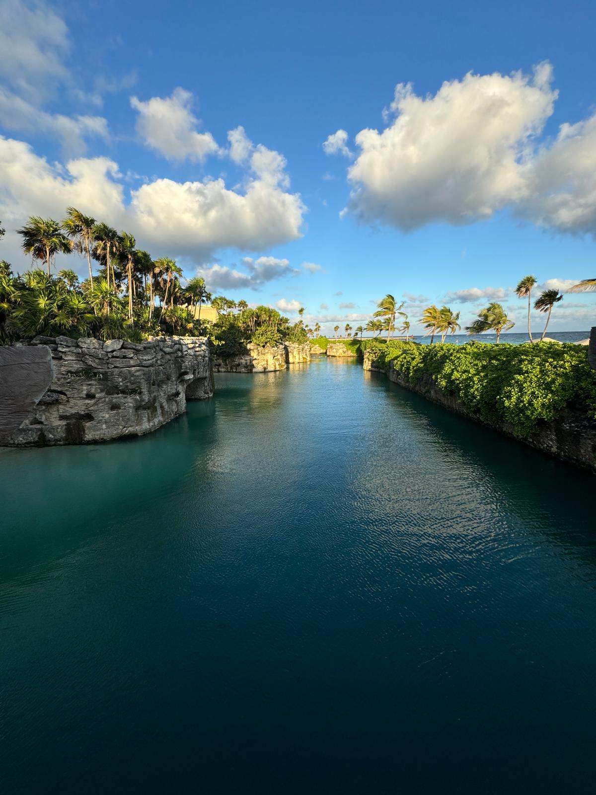 Calm blue waterway flanked by rocky cliffs and lush green palm trees under a partly cloudy blue sky.