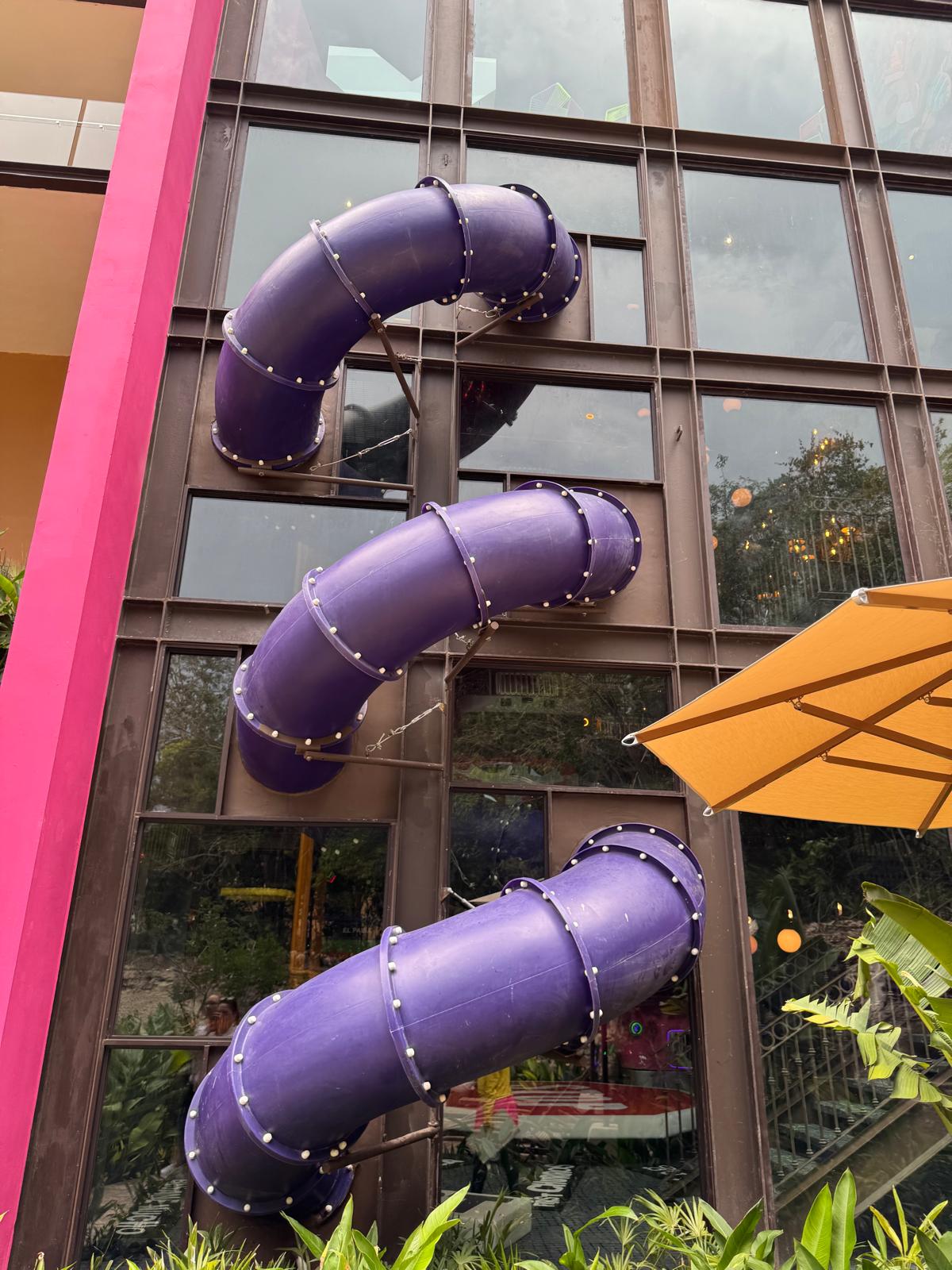 Three purple tube slides attached vertically to the exterior of a multi-story building with large windows, next to green plants and a yellow umbrella.
