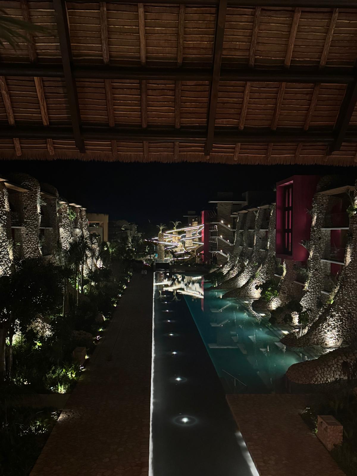 Night view of a lit outdoor pool flanked by stone buildings with illuminated palm trees and a thatched roof overhead.