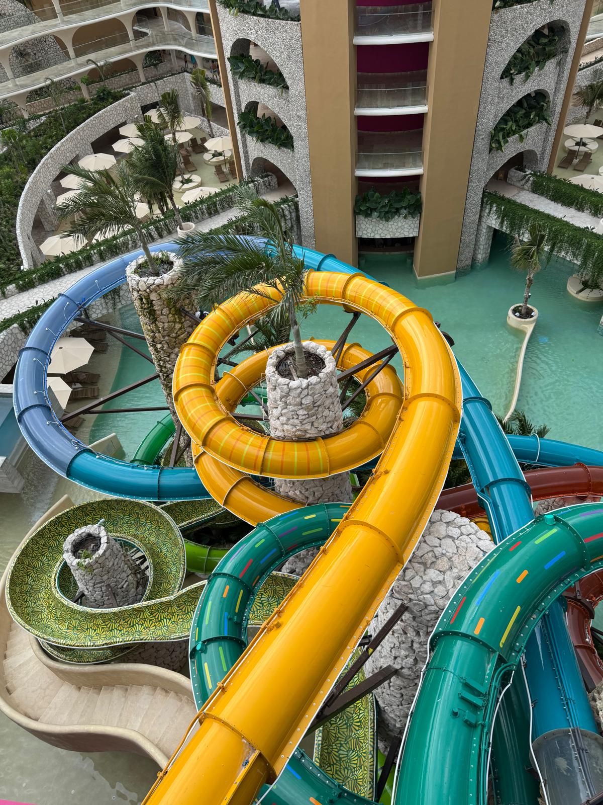 Colorful twisted water slides winding around stone pillars with palm trees, set above a pool in a resort courtyard.
