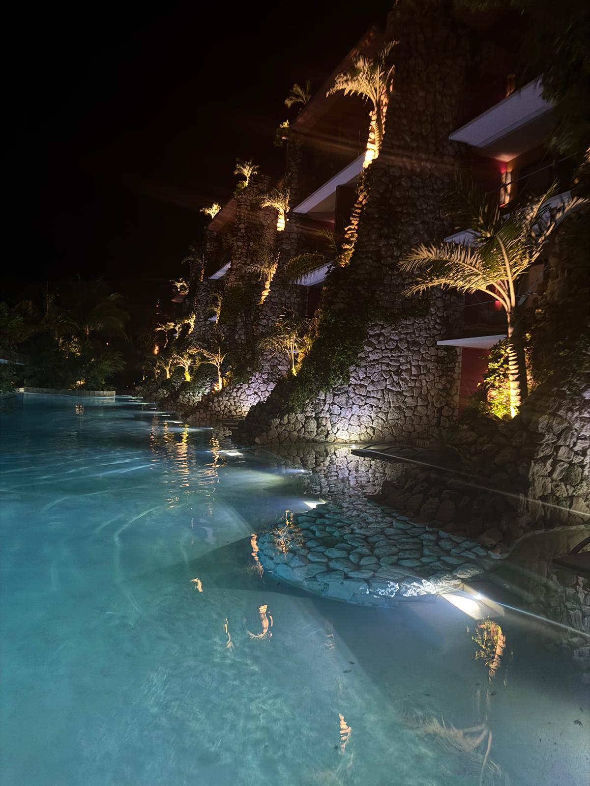 Night view of a stone building with illuminated palm trees reflected in a clear blue pool.