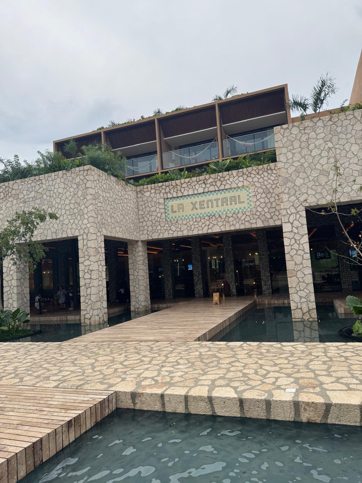 Stone building with a sign reading 'LA XENTRAL' above the entrance, surrounded by water pools and wooden walkways.