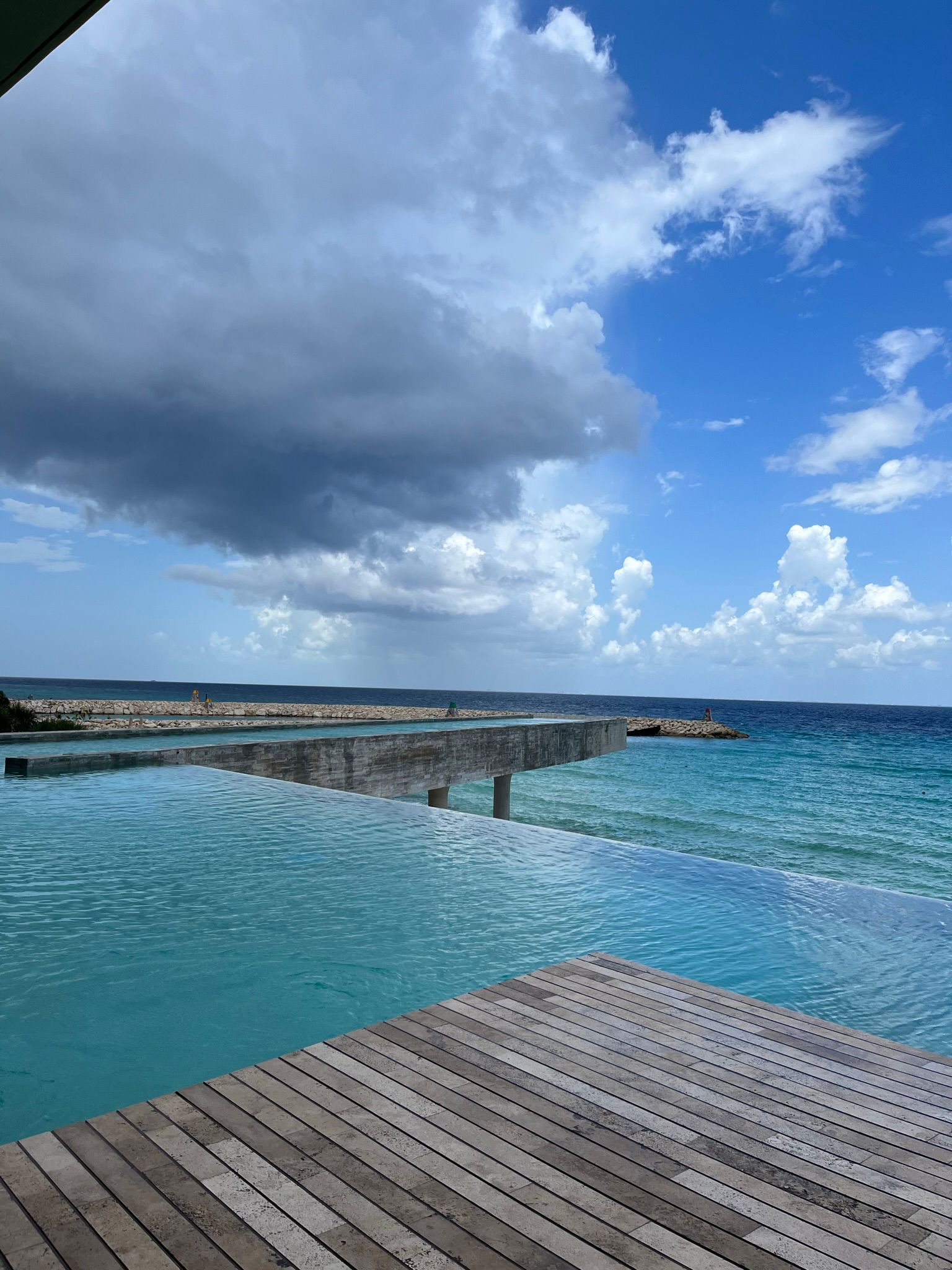 Infinity pool with wooden deck overlooking the ocean under a partly cloudy sky with dark rain clouds on the left.