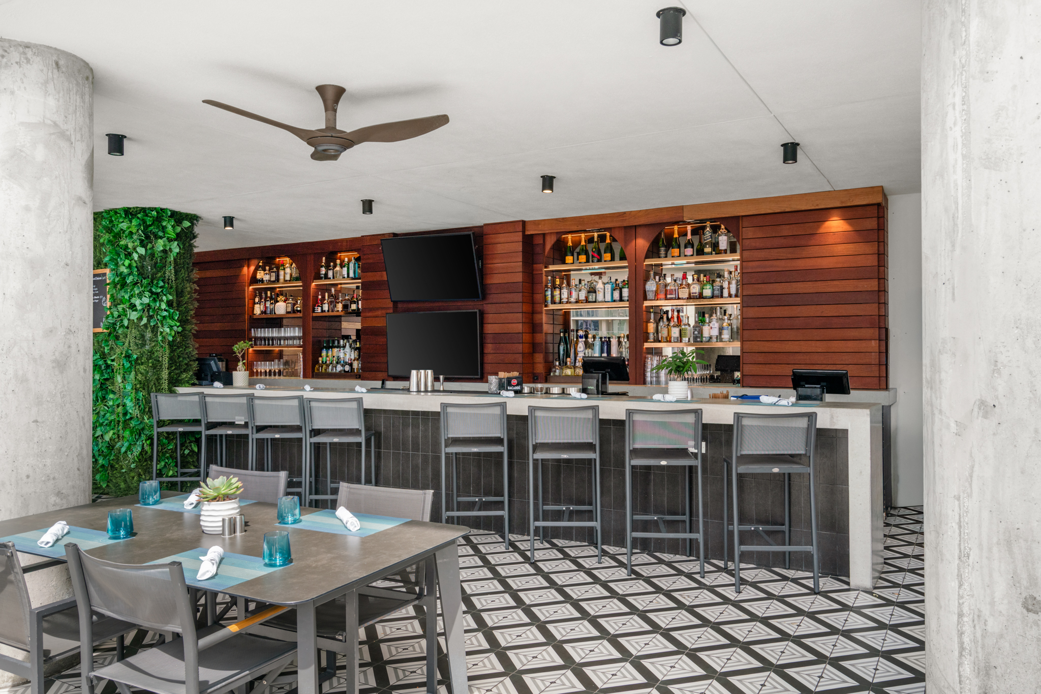 Modern bar area with high stools, liquor shelves, two wall-mounted TVs, and a dining table set with blue glasses and napkins.