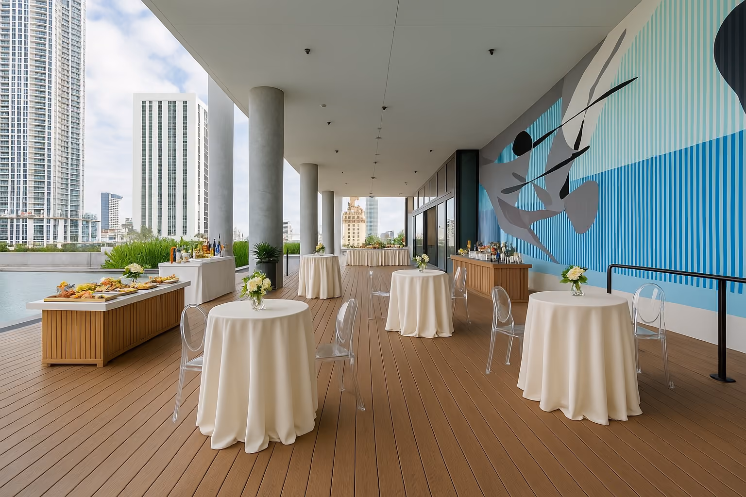 Outdoor event space with round tables covered in white linens, clear chairs, floral centerpieces, buffet and bar area, overlooking city buildings.