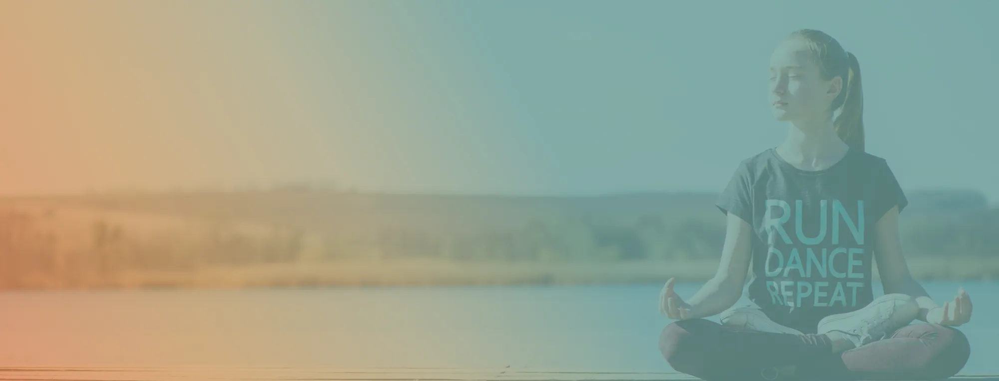 Young woman sitting cross-legged in meditation by a lake wearing a shirt that says RUN DANCE REPEAT.